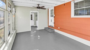 A hallway with a grey floor, white walls, and an orange accent wall. There is a ceiling fan, a window with blinds, and a door leading to another room. - Sea Breeze Retreat (North Myrtle Beach)