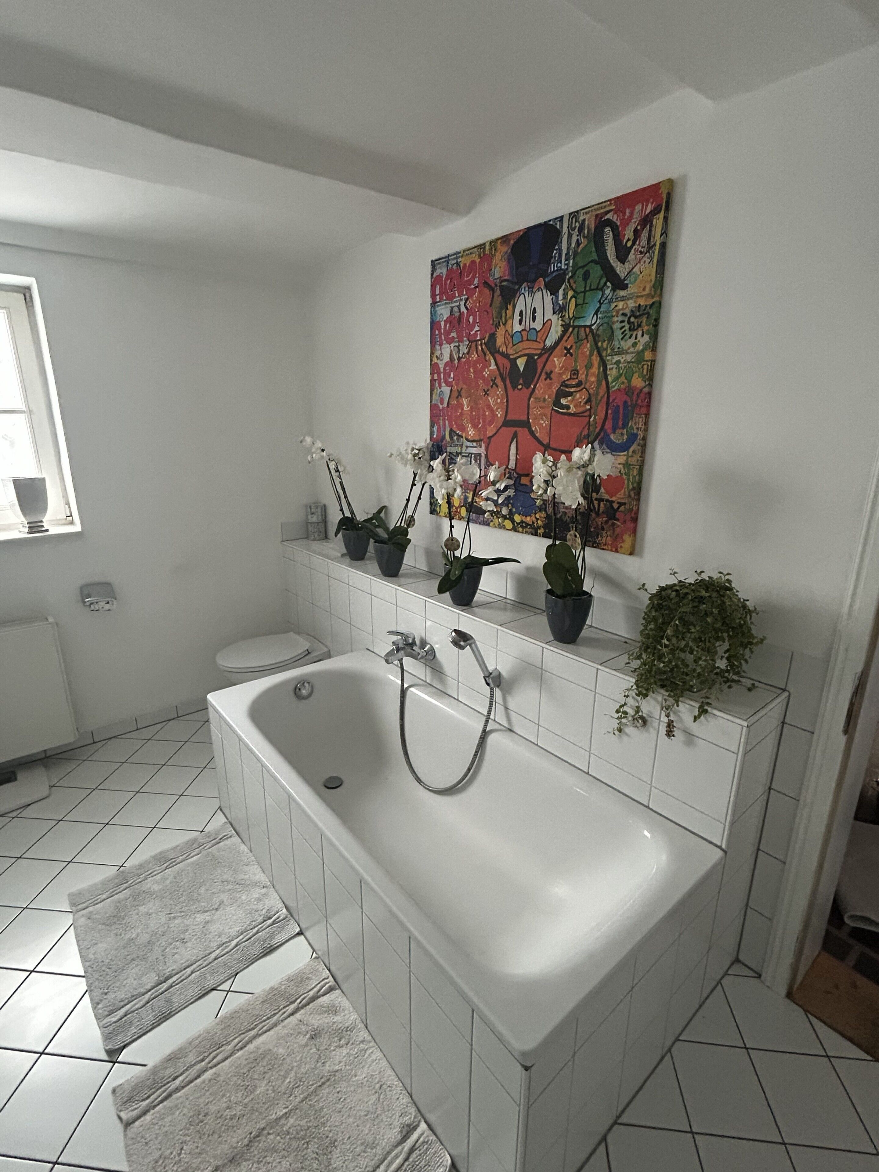 Un baño con una bañera grande, piso de baldosas blancas, un ventanal y un cuadro de colores en la pared.