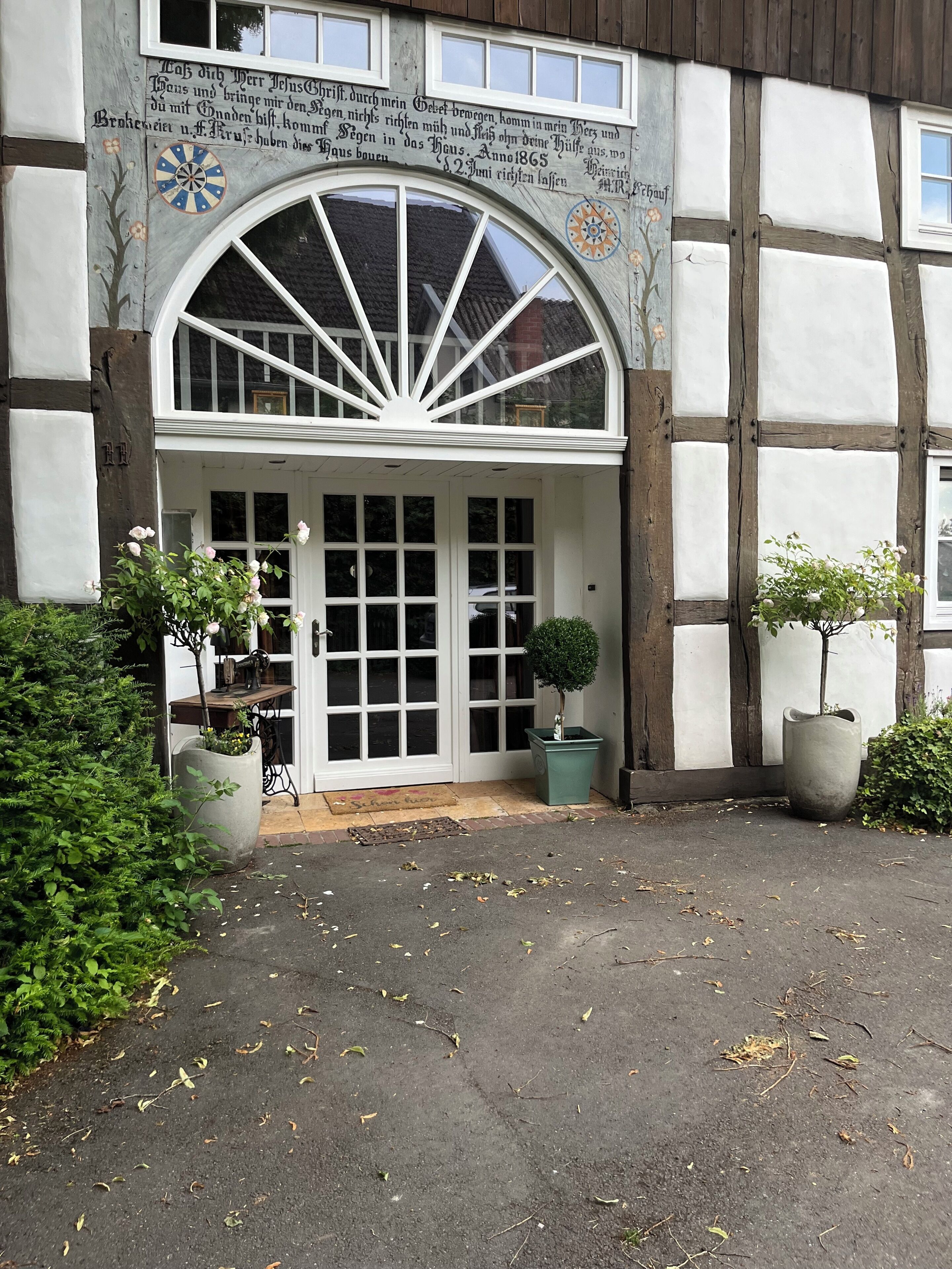A traditional half-timbered building with a large arched entrance, a door with glass panels, and a small garden area with potted plants.