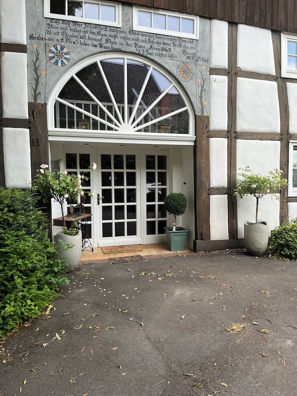 A traditional half-timbered building with a large arched entrance, a door with glass panels, and a small garden area with potted plants.