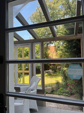 Une vue à travers une fenêtre, avec une structure en bois, une chaise et un jardin où l’on aperçoit un panneau.