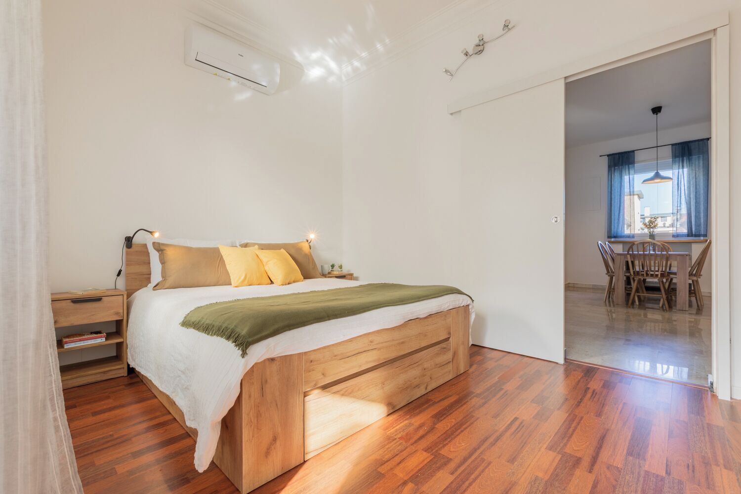 A bedroom with a wooden bed, a bedside table, a nightstand, and a window with curtains.