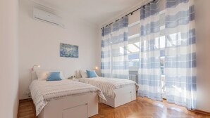 Quarto com duas camas, piso de madeira, radiador e janela com cortinas azuis.