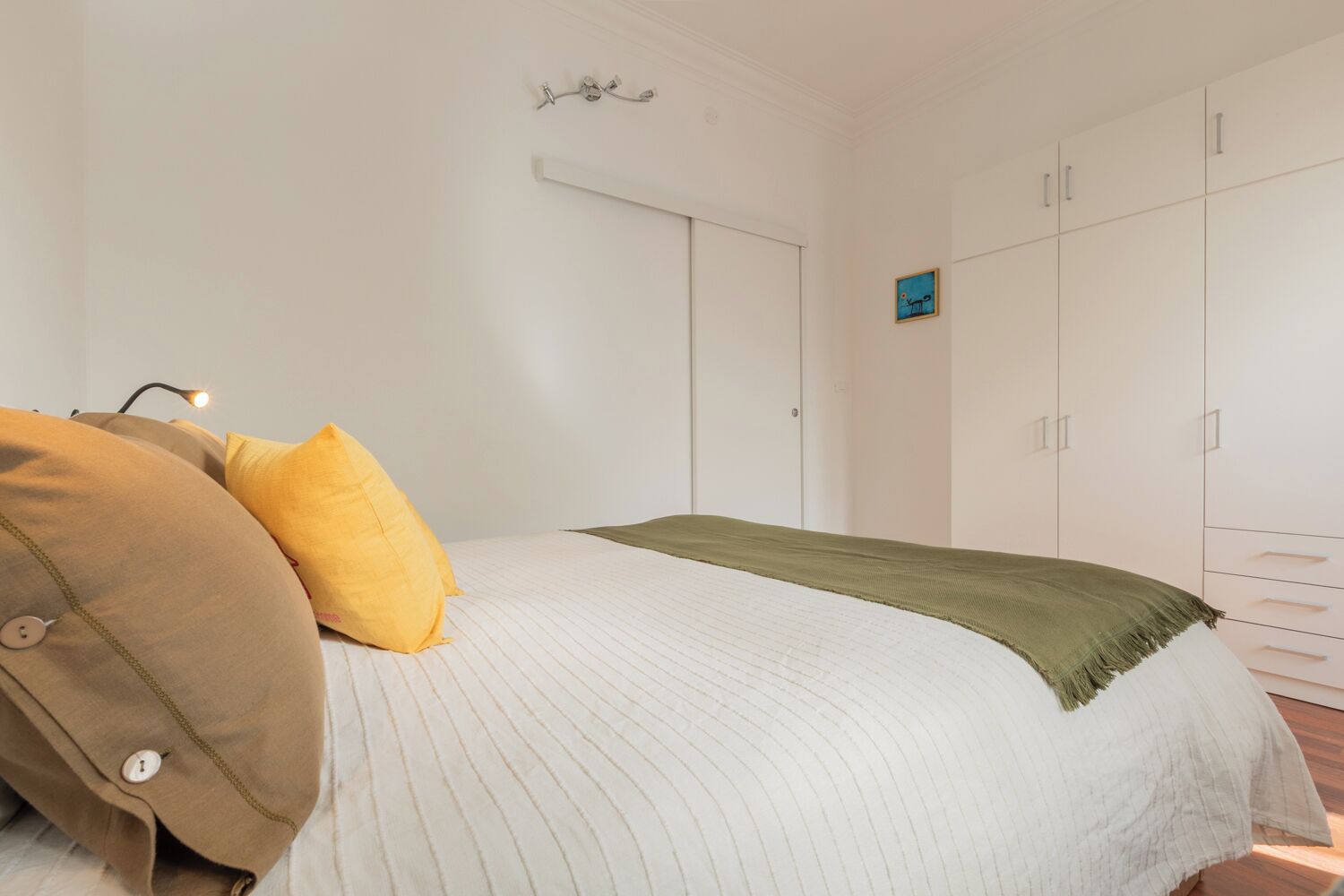 Un dormitorio con cama, almohadas, una lámpara de noche, un armario y un cuadro pequeño en la pared.