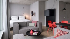 A modern hotel room with a bed, grey sofa, red chairs, a small table, and a TV.