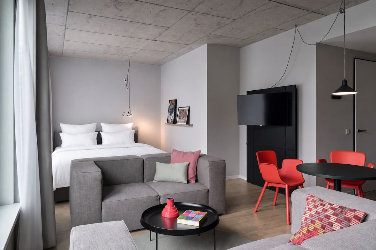 A modern hotel room with a bed, grey sofa, red chairs, a small table, and a TV.