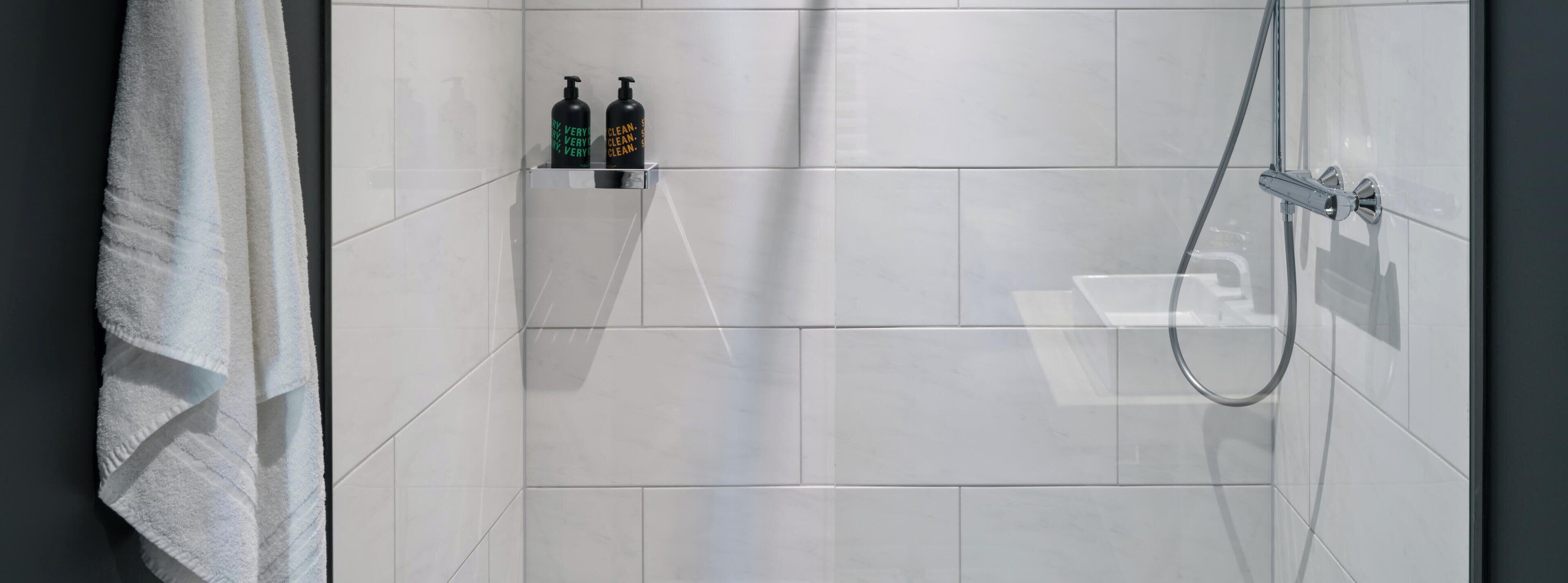 Ein Duschbereich mit weißen Fliesen, einer Glaskabine und zwei Shampoo-Flaschen auf einem Regal.