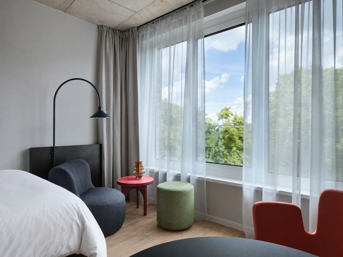 Habitación de hotel moderna con cama, escritorio, silla, lámpara y vistas a zonas verdes a través de cortinas translúcidas.