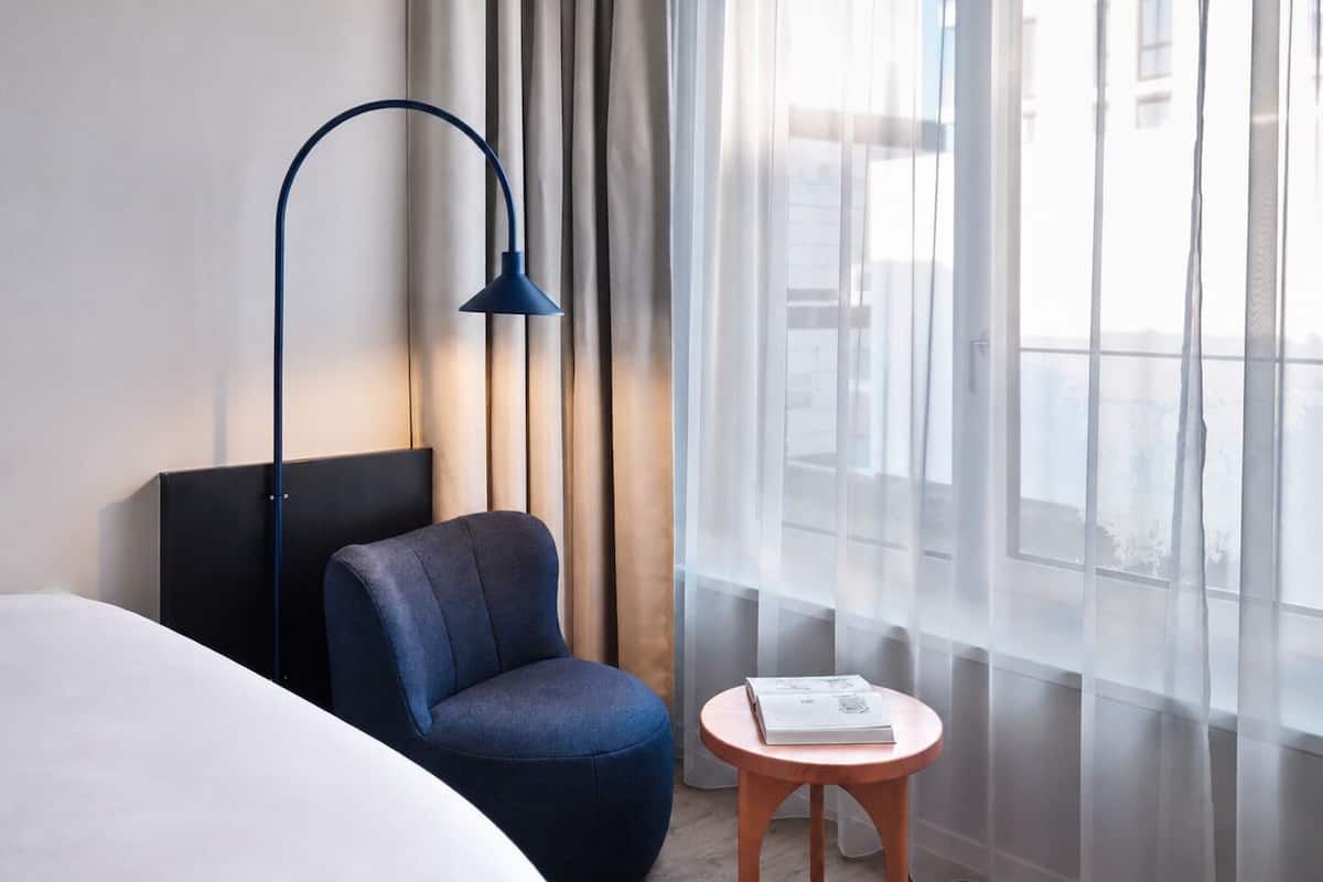 A modern hotel room with a bed, a blue chair, a small wooden table with a book, a floor lamp, and sheer curtains.