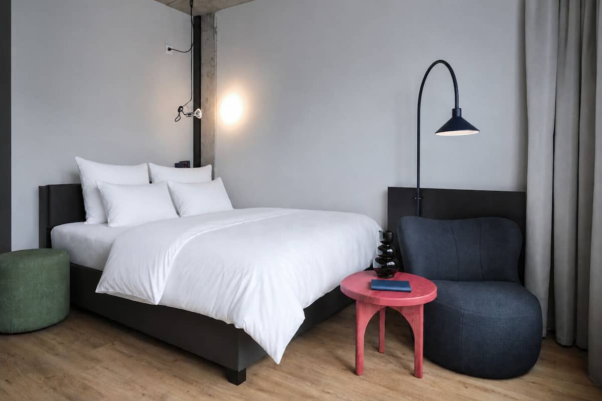 A modern hotel room with a bed, a chair, a small table, a lamp, and a wall-mounted light fixture.