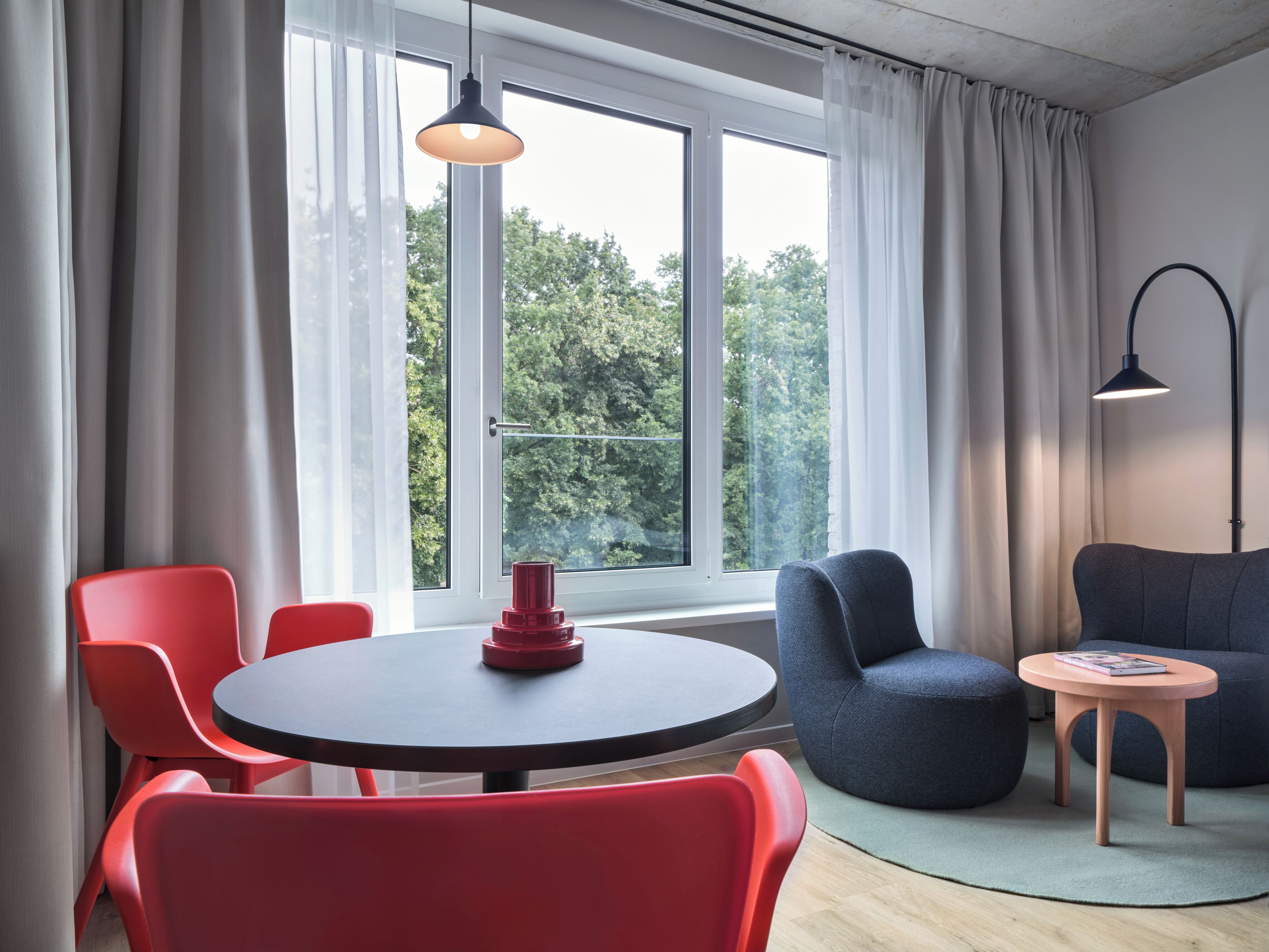 Habitació moderna amb taula rodona, cadires vermelles, sofà blau i una taula petita rodona amb una làmpada.