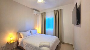 A hotel room with a bed, a TV mounted on the wall, a bedside table with a lamp, and a window with curtains. - Spacious 2BR | Apt Private Baths | Near PC Airport (Punta Cana)
