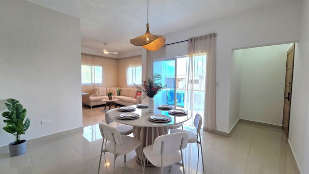 A modern dining area with a round table set for six, surrounded by white chairs. A large window provides natural light, and a ceiling fan is visible in the background. - Spacious 2BR | Apt Private Baths | Near PC Airport (Punta Cana)