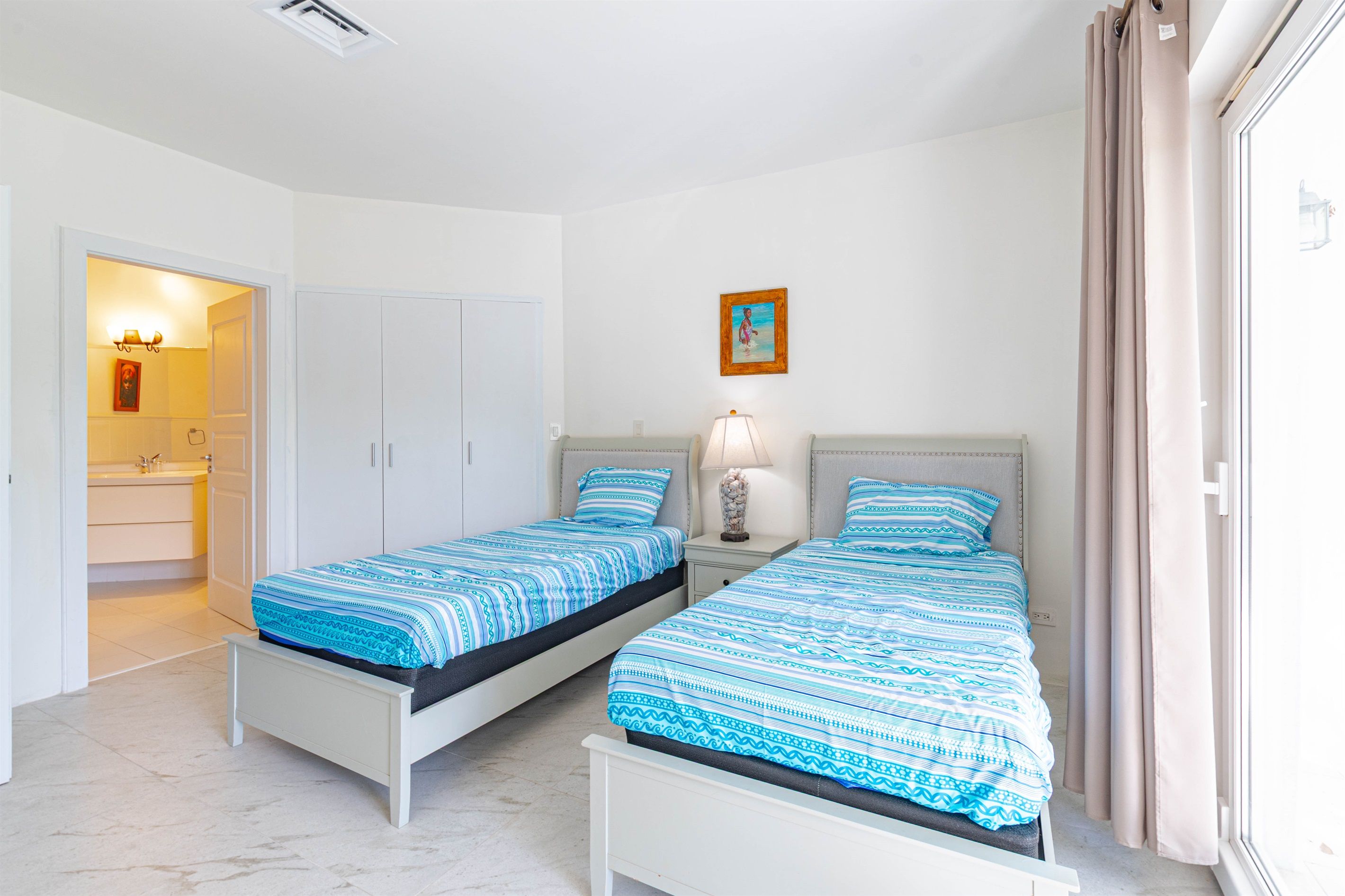 A bedroom with two beds, a nightstand, a lamp, a painting, and a bathroom visible through an open door.