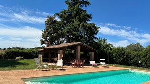 Uma piscina com um deck revestido, uma área de estar coberta ao ar livre com cadeiras e uma grande árvore ao fundo.