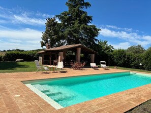 A swimming pool with a tiled deck, a covered outdoor seating area with chairs, and a large tree in the background. - Casa Petrea - Orvieto (Canale Nuovo)