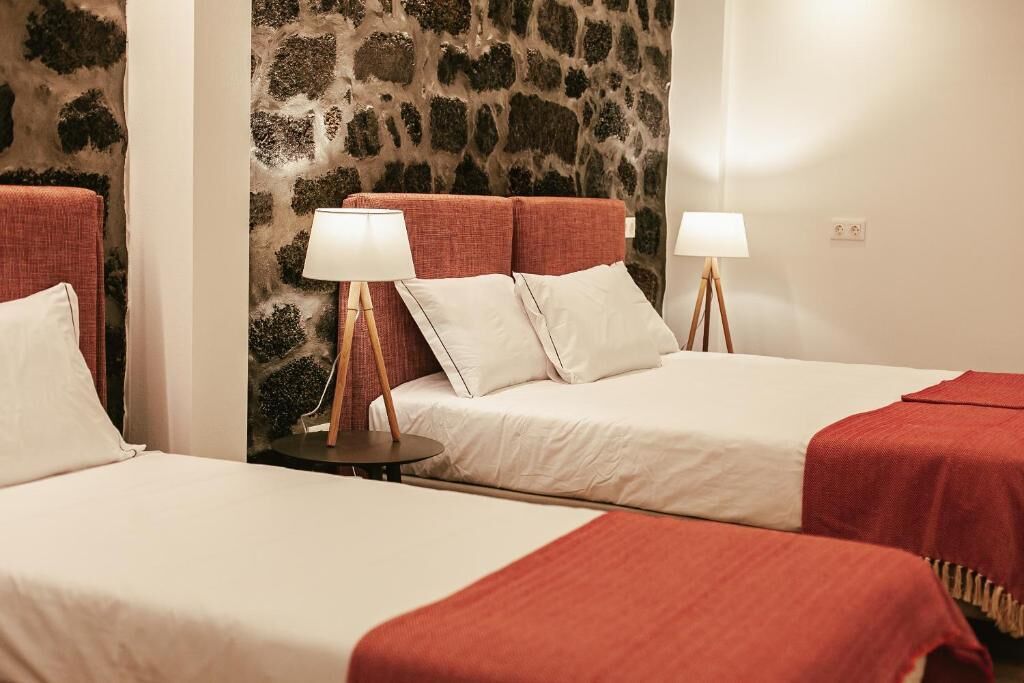 A hotel room with two beds, each with a red blanket and white pillows, a stone wall, and a small round table with a lamp.