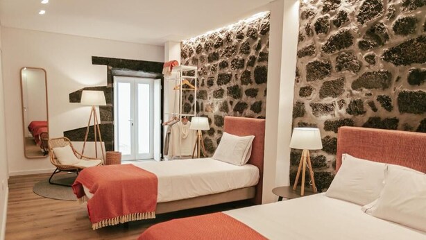 Ein Hotelzimmer mit zwei Betten, einer Steinwand, Holzboden, einem Sessel, einem Spiegel und einer Tür.