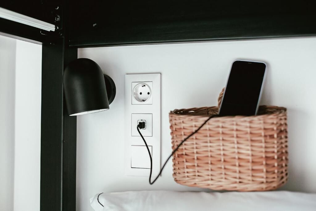 A white wall with a power outlet, a black lamp, and a wicker basket holding a smartphone.