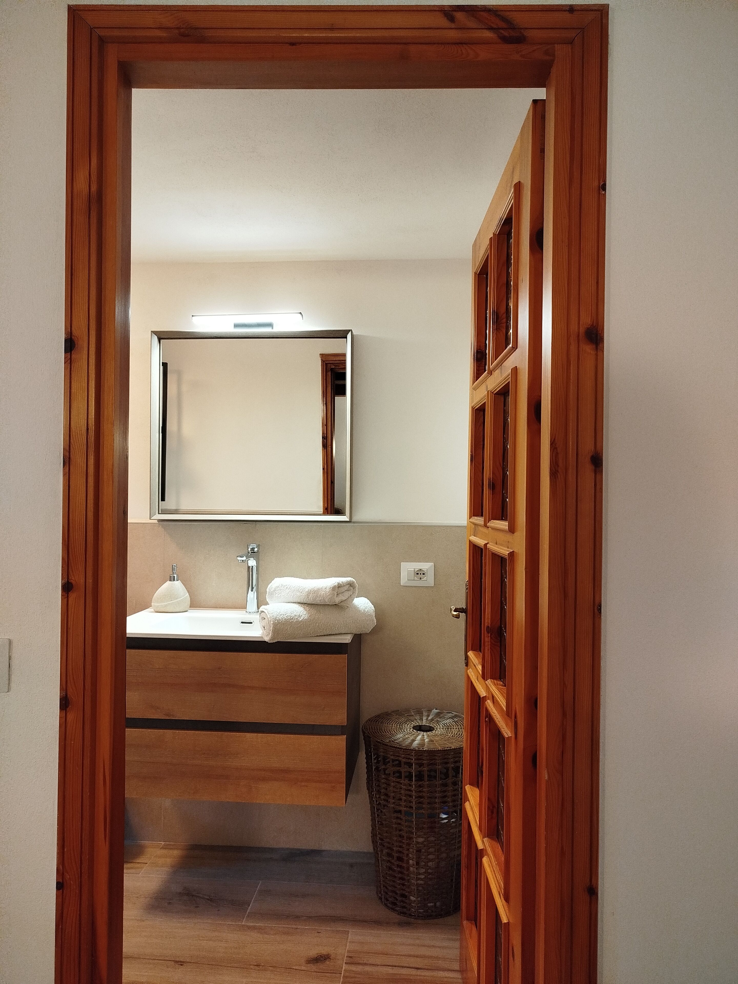 Un baño con marco de puerta de madera, un espejo sobre un lavabo, una toalla y una cesta de mimbre.