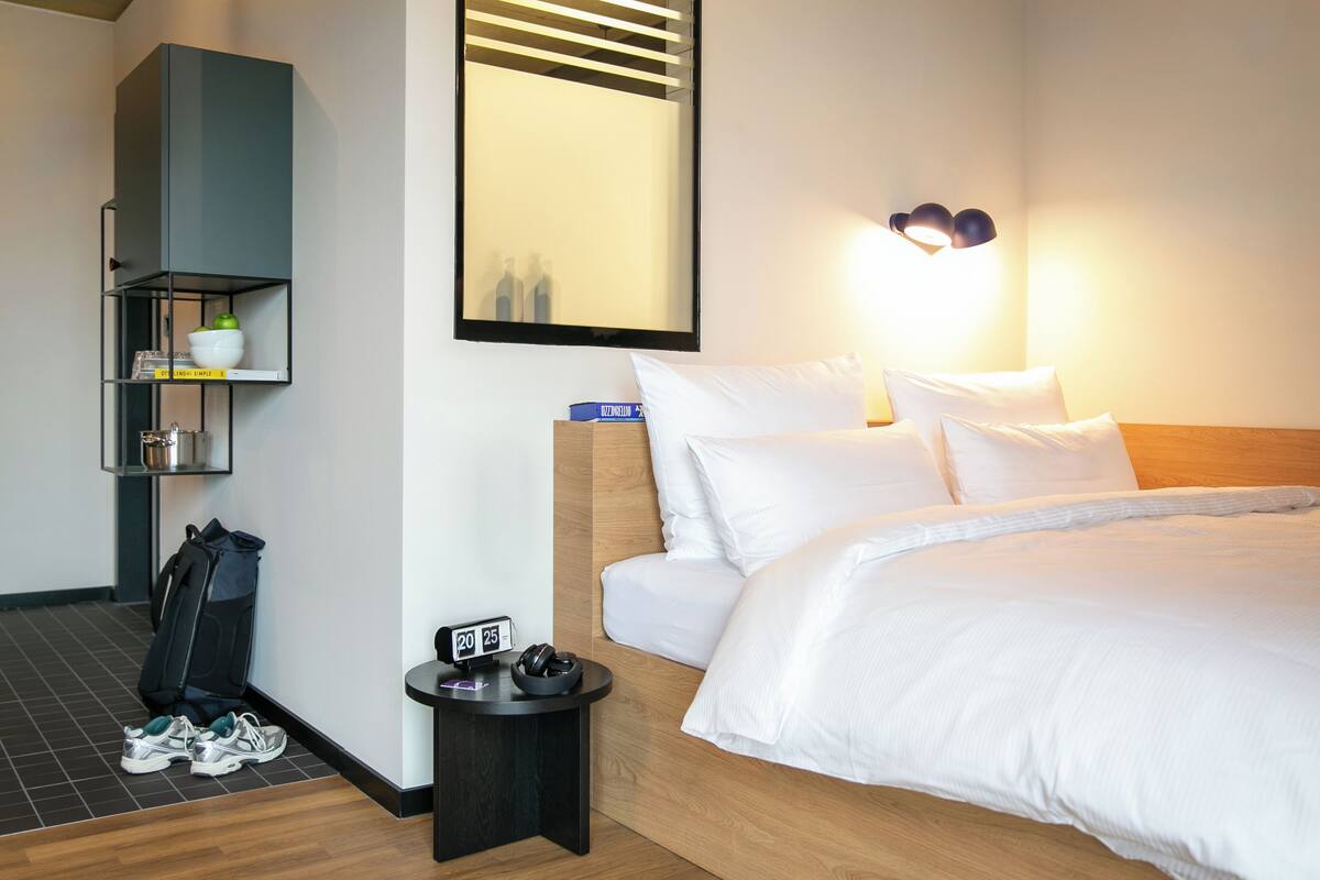A hotel room with a bed, a nightstand, a mirror, a shelf with toiletries, and a pair of shoes on the floor.