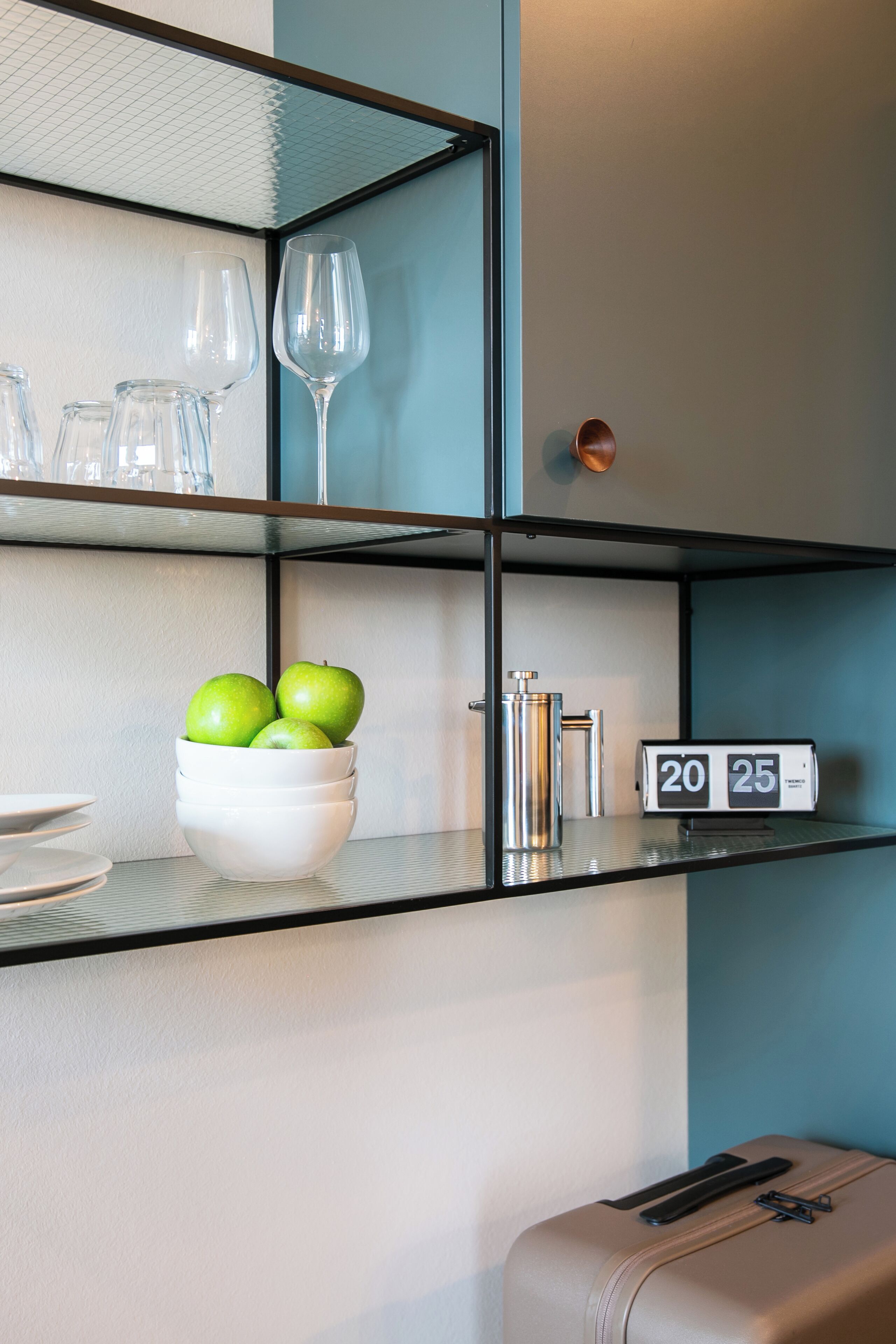 Habitación de hotel con estante de vidrio que contiene copas de vino, un cuenco con manzanas verdes, un reloj marcando las 20:25 y una maleta.