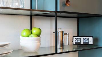 Habitación de hotel con estante de vidrio que contiene copas de vino, un cuenco con manzanas verdes, un reloj marcando las 20:25 y una maleta.