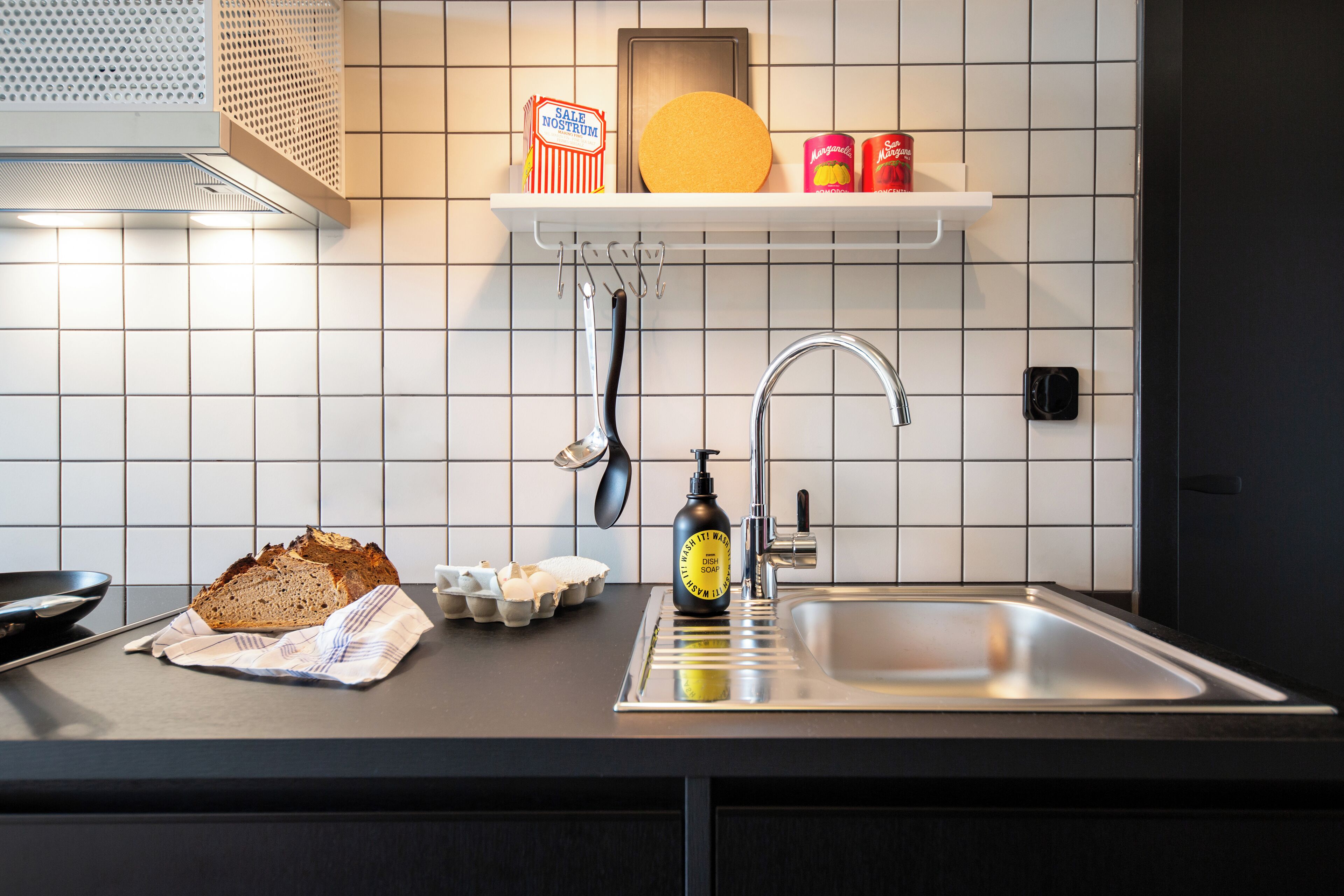 A kitchen with a stainless steel sink, a loaf of bread, eggs, a bottle of soap, and a shelf with a yellow plate and canned goods.