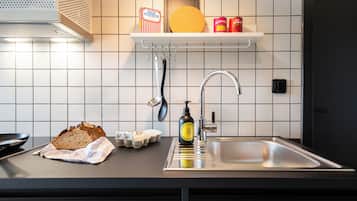 Et køkken med en stålvask, et brød, æg, en sæbeflaske og en hylde med en gul tallerken samt dåsemad.