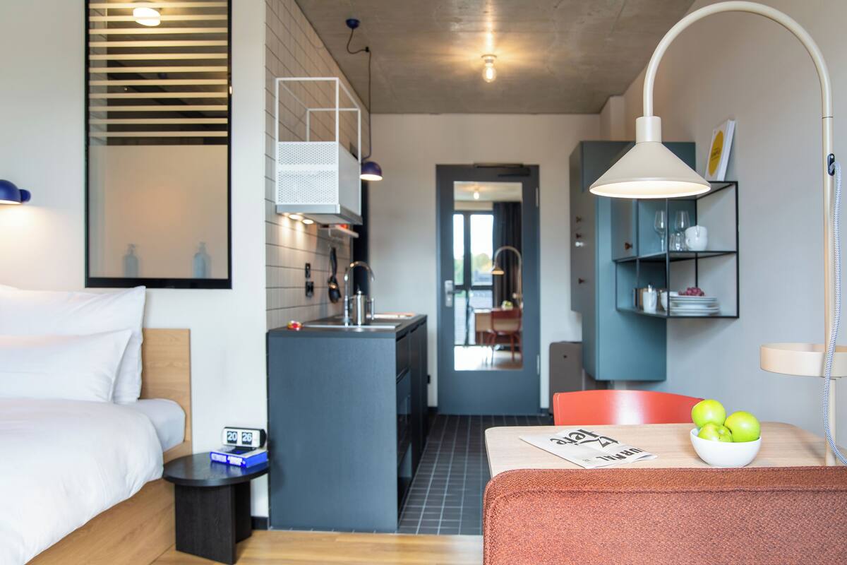 A modern hotel room with a bed, a small kitchenette, a dining area, and a bathroom.