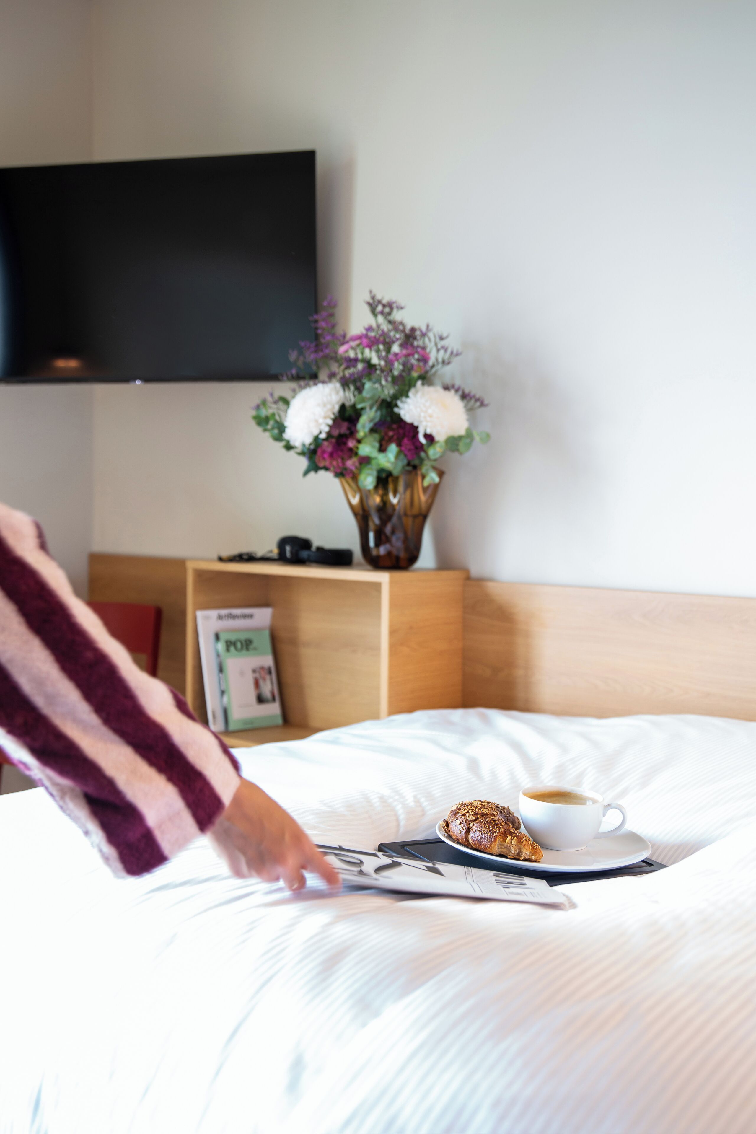 Een netjes opgemaakt bed met een kop koffie en een croissant, een bloevas en een flatscreen televisie aan de muur.
