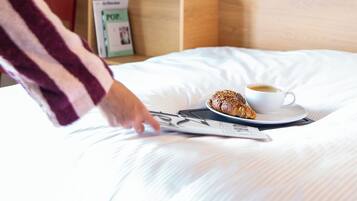 Een netjes opgemaakt bed met een kop koffie en een croissant, een bloevas en een flatscreen televisie aan de muur.