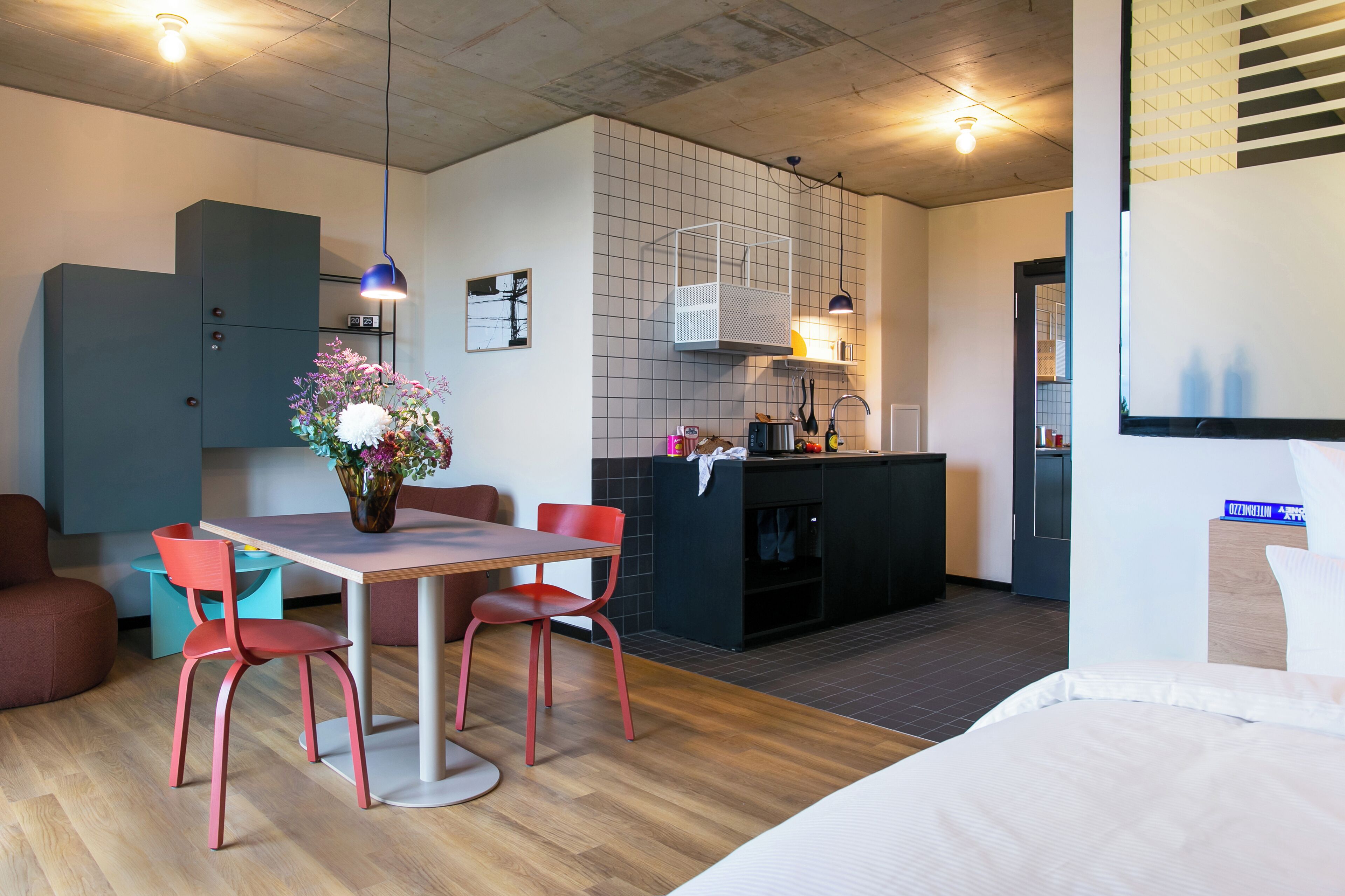 A modern hotel room with a wooden dining table, red chairs, a small kitchen area, and a bed.