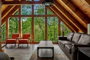 A spacious loft with a high ceiling, large windows offering a view of lush greenery, a leather sofa, and two armchairs. - The Mountain Estate by Ghosal Luxury Lodging (Gatlinburg)