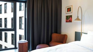 A hotel room with a bed, a chair, a side table, a lamp, and a view of buildings through a window with curtains.