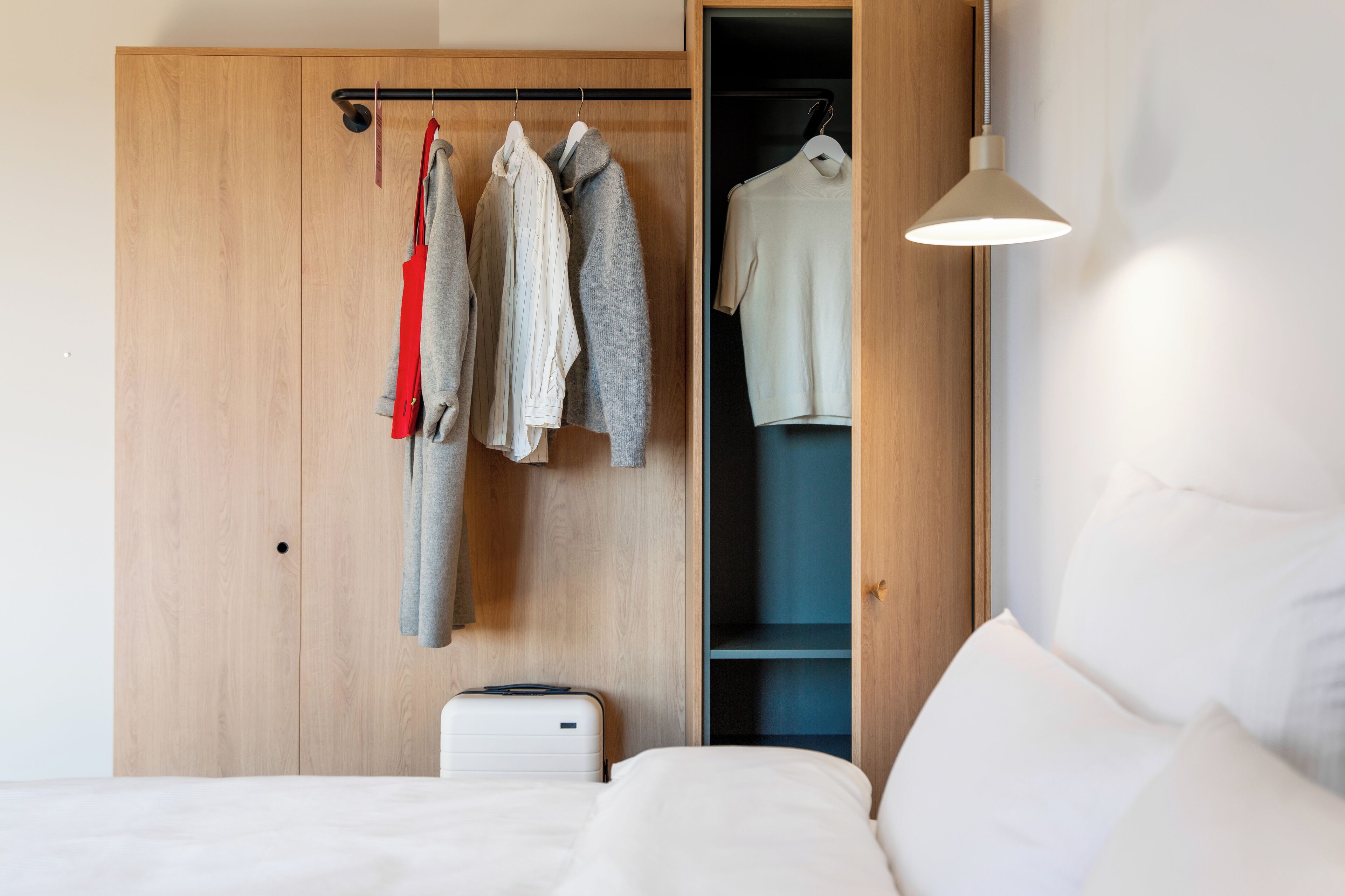 A bedroom with a wooden wardrobe, a hanging rack with clothes, a bed with white bedding, and a bedside table.
