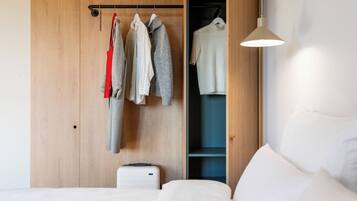 A bedroom with a wooden wardrobe, a hanging rack with clothes, a bed with white bedding, and a bedside table.