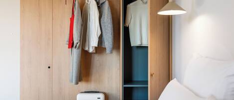 A bedroom with a wooden wardrobe, a hanging rack with clothes, a bed with white bedding, and a bedside table.