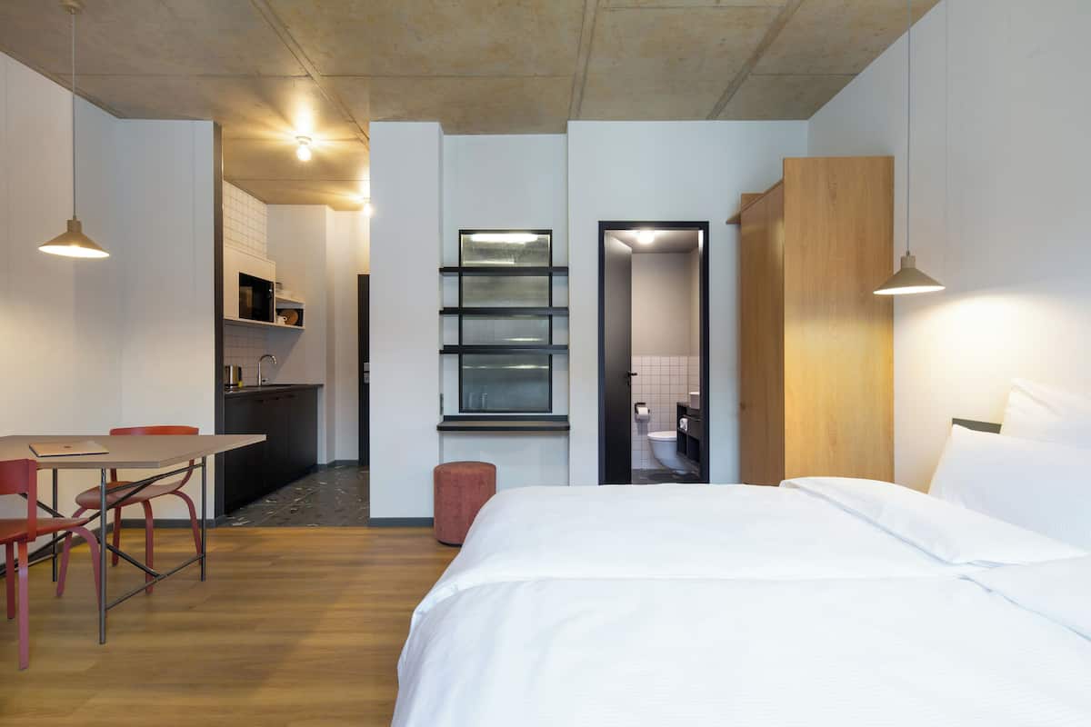 A modern hotel room with a wooden floor, a bed with white linens, a dining table with chairs, and a kitchenette in the background.