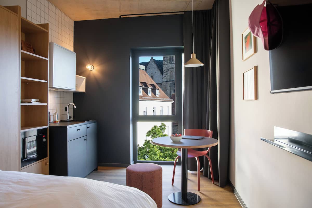 A modern hotel room with a bed, a small kitchenette, a dining area with a table and chairs, and a large window offering a view of buildings and trees.