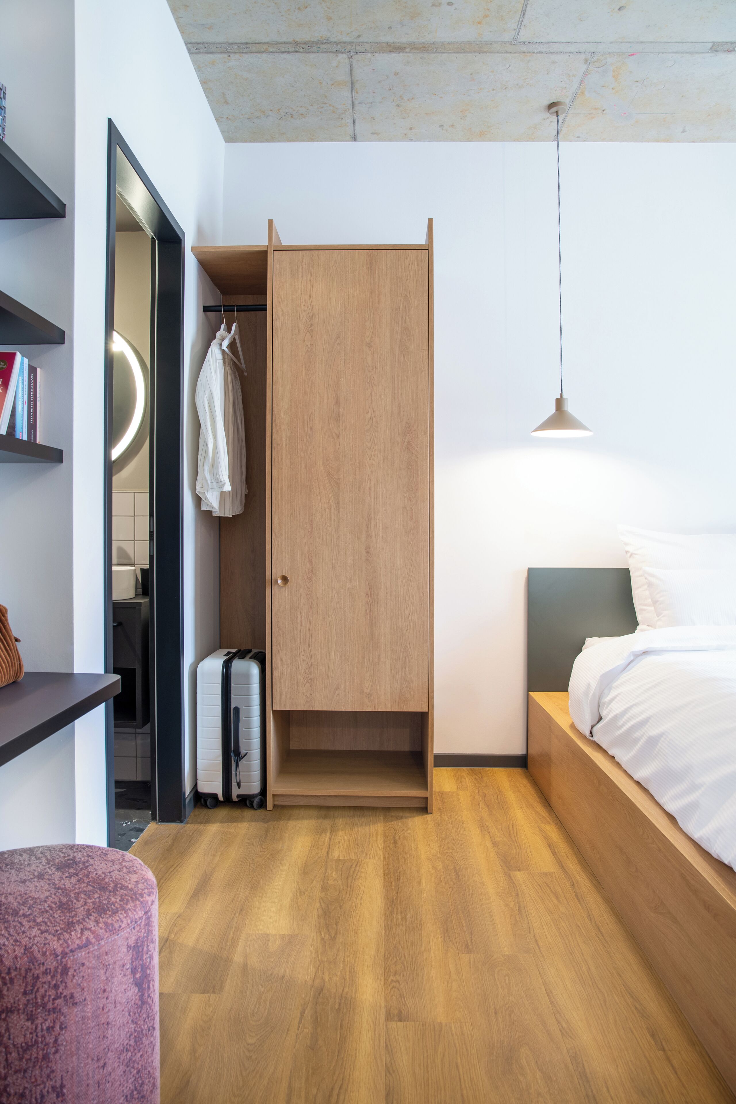 Une chambre à coucher de taille réduite, comprenant un lit, une armoire, un petit coin salon et une valise.
