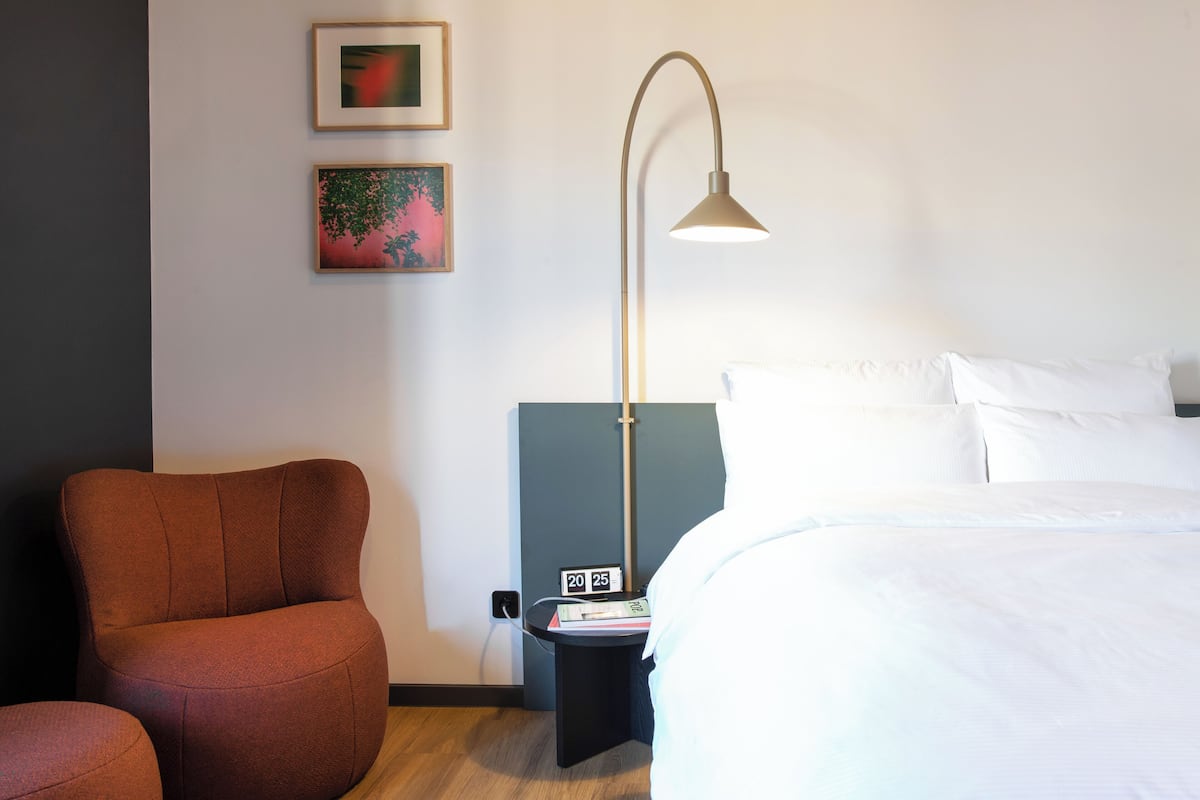 A hotel room with a bed, a red armchair, a nightstand with a lamp, and two framed pictures on the wall.