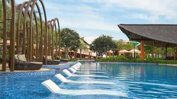 Un área de piscina con sillones de descanso y sombrillas, rodeada de palmeras y una zona de estar cubierta.