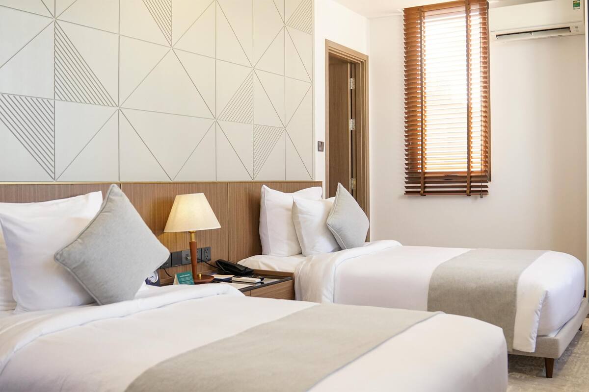 Une chambre d’hôtel avec deux lits, un mur orné d’un motif géométrique, une tête de lit en bois, une table de chevet avec une lampe et une fenêtre avec des stores.