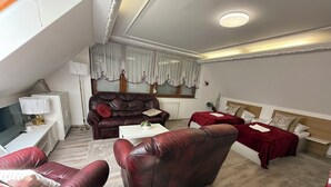 Ein modernes Wohnzimmer mit einer Ledersofa, einem weißen Couchtisch und einem roten Sofa. Ein Fernseher steht auf einer weißen Konsole, und ein Fenster mit transparenten Vorhängen.