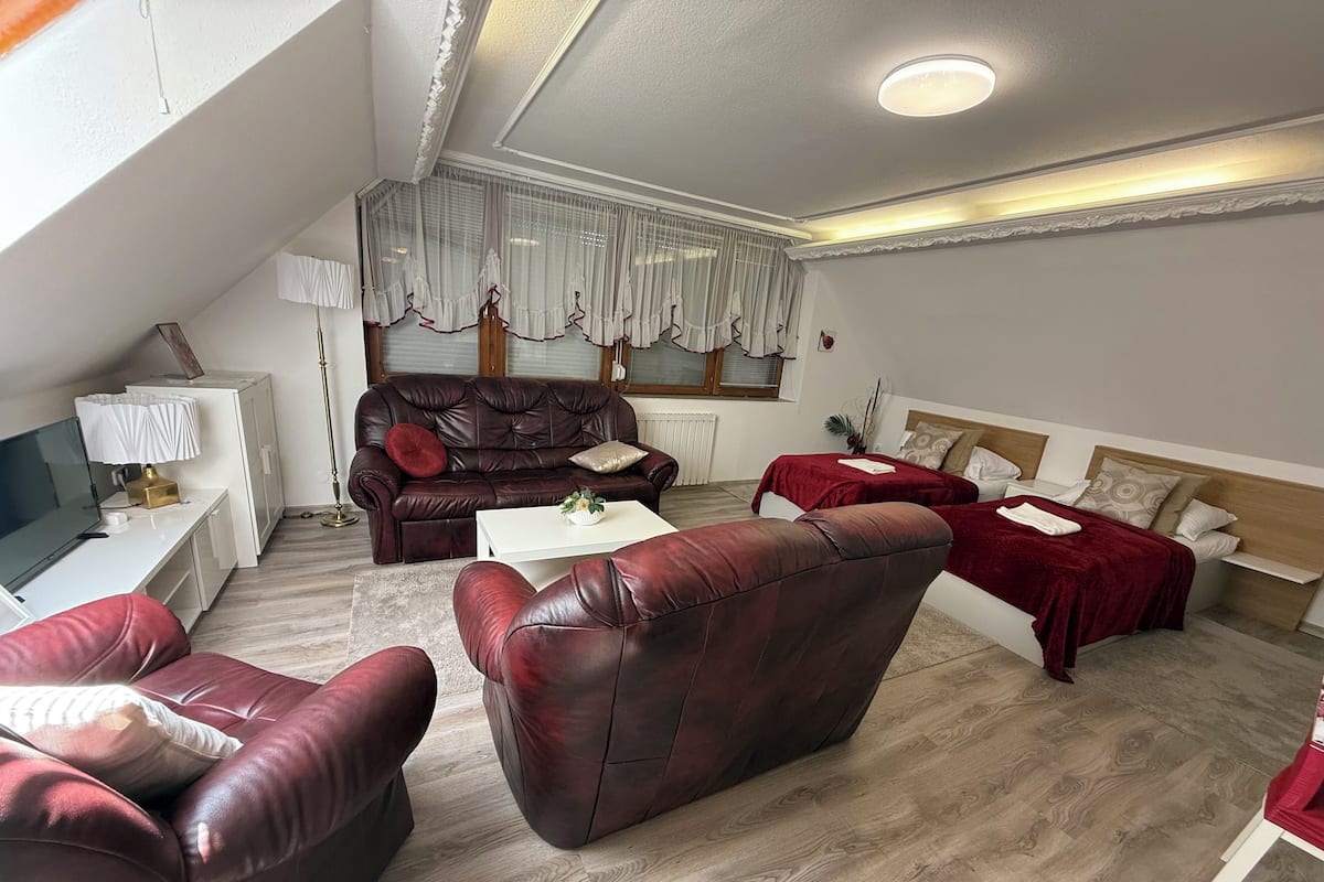 Ein modernes Wohnzimmer mit einer Ledersofa, einem weißen Couchtisch und einem roten Sofa. Ein Fernseher steht auf einer weißen Konsole, und ein Fenster mit transparenten Vorhängen.