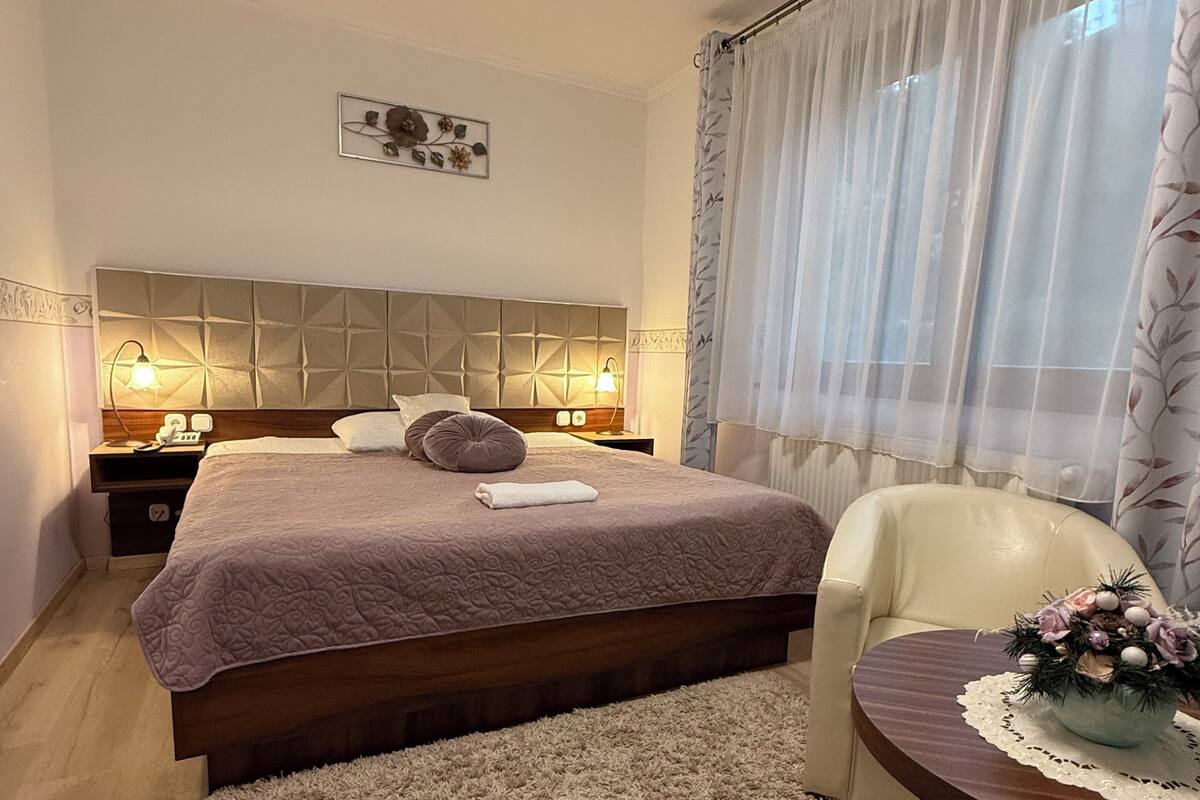 Ein ordentlich eingerichtetes Schlafzimmer mit einem Bett, Nachttischlampen, einem Sessel und einem kleinen Tisch mit einer Dekoration.