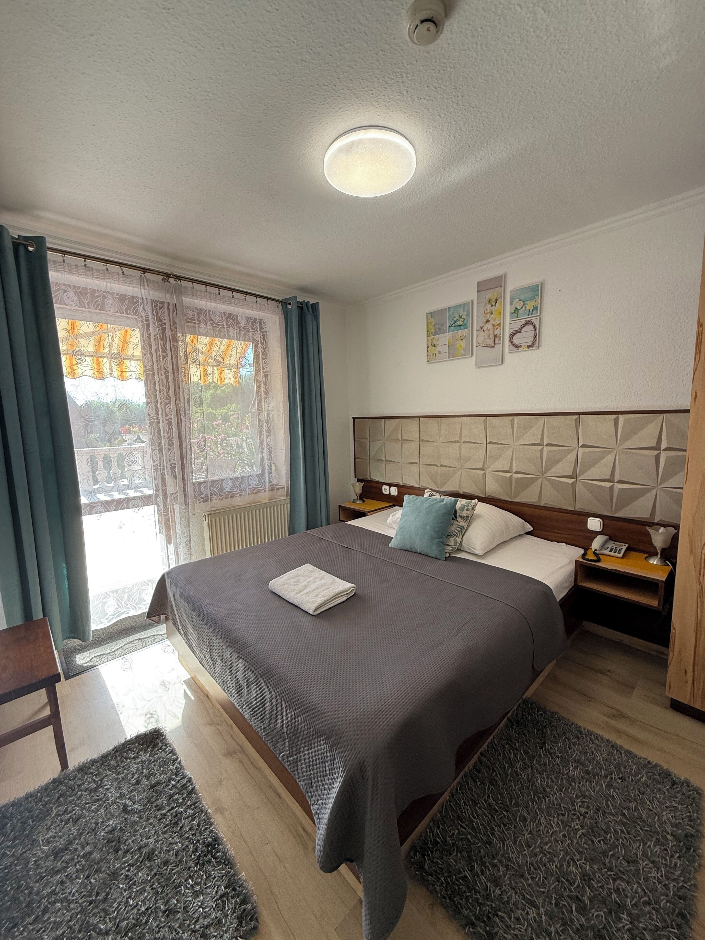 A hotel room with a large bed, a bedside table with a lamp, a nightstand, a window with curtains, and a ceiling light.