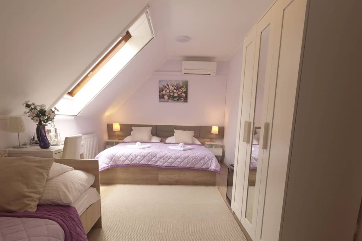 Ein Schlafzimmer mit schrägen Wänden, einem Bett mit lila Bettdecke, einem Nachttisch, einer Lampe, einer Vase mit Blumen und einem Kleiderschrank.