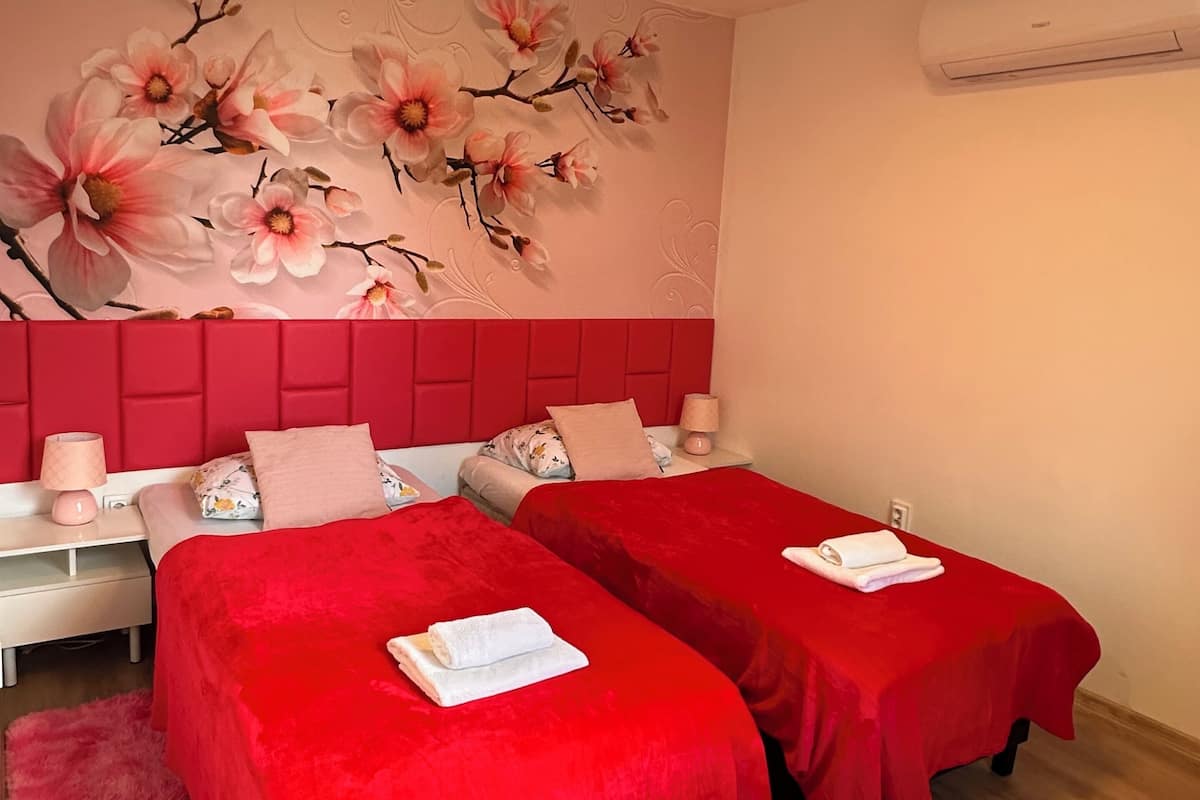 Ein Hotelzimmer mit zwei Betten, bezogen mit roten Laken, einem floralen Wandbild, einem roten Kopfteil, Nachttischen mit Lampen und einer Klimaanlage an der Wand.
